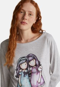 Rothaarige Frau mit Sommersprossen, die ein graues Hemd trägt, auf dem eine farbenfrohe Illustration von zwei nebeneinander stehenden Mädchen in Pastellkleidern abgebildet ist.