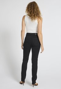 Käharate juustega naine, kes kannab valget varrukateta topi, musti kitsaid pükse ja pruune kontsakingi, seisab valgel taustal seljaga vaataja poole.