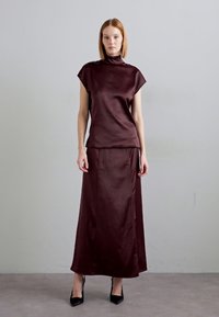 Burgundska satenasta majica z visokim ovratnikom in kratkimi rokavi, v kompletu z dolgim satenastim krilom v enaki barvi. Model nosi črne špicaste petke.
