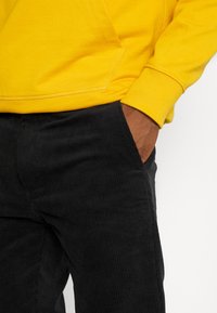 Gul sweatshirt med känguruficka, ribbade ärmslut; svarta kordbyxor med breda, vertikala ribbor och sidoficka synlig.