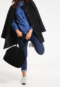 Svart kappa över en denimskjorta, tillsammans med blå jeans och vita sneakers. Har också en svart tygväska med rundade kanter, som hålls i handen.