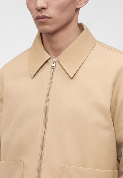 Homme portant une veste beige avec un col pointu et une fermeture éclair devant, montrant le haut du torse et une expression faciale neutre.