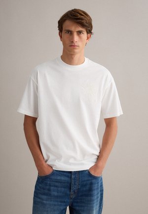 Giovane uomo con capelli castani che indossa una semplice maglietta bianca e jeans blu, in piedi con le mani in tasca contro uno sfondo neutro.