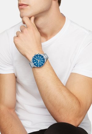 Montre chronographe bleue avec bracelet en métal argenté, dotée d'un cadran rond, d'indicateurs horaires blancs et de plusieurs cadrans pour diverses fonctions.