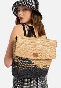 Zaino intrecciato in paglia con una sfumatura di beige naturale e blu scuro; presenta una patta pieghevole e un piccolo logo in pelle come accent. Spallacci neri.