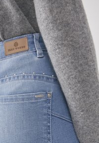 Jeans de mezclilla azul claro con una etiqueta de marca de cuero, sutiles tachuelas plateadas en el bolsillo trasero y un suéter de punto gris parcialmente visible.