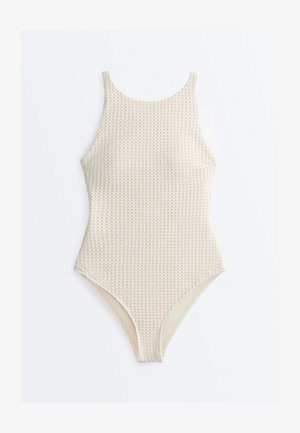 Krém színű, texturált egyrészes fürdőruha, magas nyakkal és mérsékelt lábszárvágással, fehér háttéren elhelyezve.