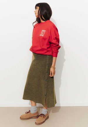 Sweatshirt rouge avec texte, jupe en tricot côtelé verte avec boutons sur le côté, chaussettes grises, chaussures beiges avec lacets marron, le tout sur fond blanc.
