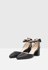 Scarpe nere in pelle con tacco alto, punte affusolate, cinturini alla caviglia e tacco a blocco. Caratterizzate da una morbida fodera interna e dettaglio a fiocco sul retro.