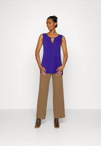 Top roxo sem mangas com detalhe em fenda, combinado com calças largas de cintura alta castanhas e botas de tornozelo castanhas sobre um fundo liso.