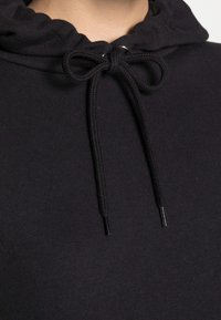 Zwarte hoodie van een textiele stof; voorzien van een trekkoord met zwarte aglets en een gathering in de capuchon. Close-up weergave van de kraag en ontwerpdetails.