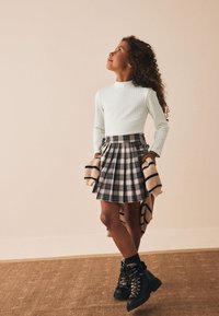 Top de manga larga de canalé verde claro, falda plisada a cuadros en blanco y negro, y botas negras con cordones. La modelo sostiene un suéter beige a rayas.