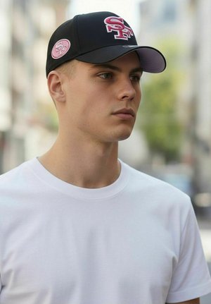 Young man wearing white t-shirt and black baseball cap with "SF" logo, facing right on an urban street background.