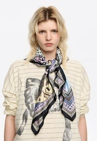 Blond persoon in een gestreepte beige shirt met opgerolde mouwen en een gekleurde, patronen sjaal om de nek, tegen een witte achtergrond.