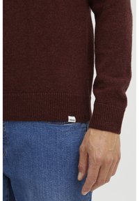 Burgundy wool sweater with ribbed cuffs and hem. Label at the side seam. Worn with blue denim jeans, showcasing a casual fit.