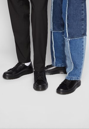 Black lace-up sneakers with round toes and rubber soles, worn with black and patchwork denim pants featuring frayed edges.