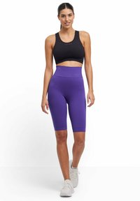 Zwarte sportbeha en paarse textuur fietsbroek, hoog in de taille, naadloze stof, gedragen met witte sportschoenen.