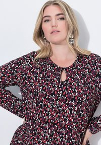 Langemouwen blouse met een ronde halslijn en een sleutelgatdetails, bedekt met een patroon van rode, witte en paarse diamanten op een zwarte achtergrond.