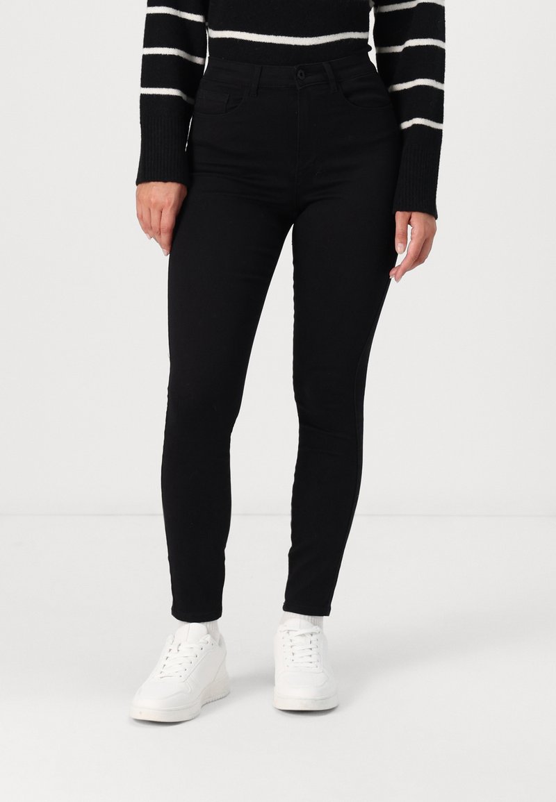 Personne portant un jean slim noir, un pull rayé noir et blanc, des chaussettes blanches et des baskets blanches, debout sur un fond blanc.
