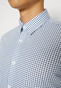 Homme portant une chemise blanche boutonnée avec un motif à carreaux bleus et un col pointu, visible du cou à la poitrine.