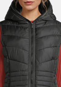 Vrouw in een zwart gewatteerd vest met capuchon en een voorrits over een lange mouw ribbelshirt in het rood, cropped bij de nek en torso.