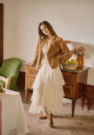 Femme appuyée sur un buffet en bois avec un panier de citrons, portant une veste marron et une jupe blanche superposée dans une pièce confortable avec un fauteuil vert.
