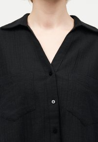 Κοντινό πλάνο ενός ατόμου που φοράει μαύρο, υφασμάτινο πουκάμισο με κουμπιά, γιακά και δύο τσέπες στο στήθος, εμφανίζοντας τον λαιμό και το άνω μέρος του στήθους.