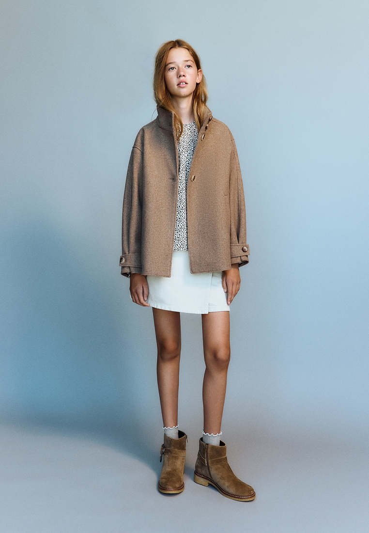 Jeune fille portant un manteau marron, un haut à motifs, une jupe blanche et des bottines marron, debout devant un fond uni bleu clair.