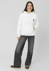 Jeune femme souriante portant un sweat à capuche blanc avec un petit motif de main, un jean ample de couleur foncée et des baskets marron, debout sur un fond uni.