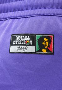 Μπαλώματα σε μωβ ύφασμα με το κείμενο "Το ποδόσφαιρο είναι ελευθερία", ένα πολύχρωμο στρογγυλό λογότυπο, και μια στυλιζαρισμένη πορτρέτο σε κόκκινα, κίτρινα και πράσινα χρώματα.
