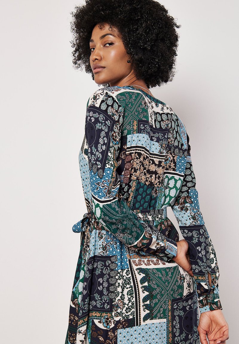 Pestré šaty se vzorem, které obsahují různé designy v zelené, modré a námořnické barvě, s dlouhými rukávy, páskem na zavázání v pase a volným střihem.