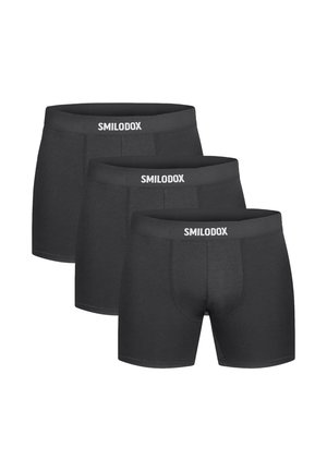 Muskeløs mand med kort hår iført sorte boxershorts og hvide ankelsokker, der står mod en ensfarvet lys baggrund.