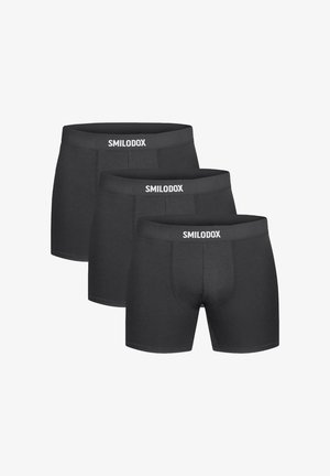 Gespierde man met kort haar, die zwarte boxershort en witte enkelsokken draagt, staand tegen een effen lichte achtergrond.