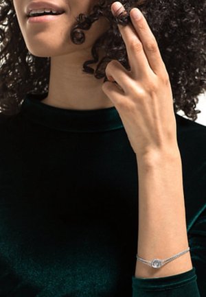 Femme aux cheveux bouclés portant un haut en velours vert foncé, enroulant une boucle avec ses doigts, portant un bracelet en argent au poignet.