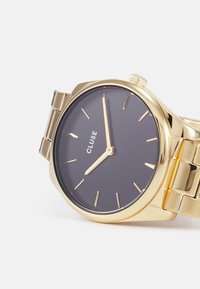 Gold-tone wristwatch with a dark gray dial. Features minimalistic hour markers and hands, metal bracelet, and a rounded case.