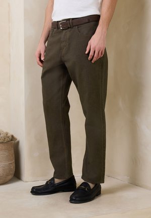 Mann trägt dunkelbraune Hose, schwarze Loafers und weißes Hemd, steht drinnen nahe einer beigen Wand und einem dekorativen Korb.