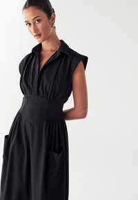 Ženska s temnimi lasmi v črni obleki brez rokavov z velikimi žepi, ki stoji ob enostavni svetli podlagi in nosi zlate uhane.