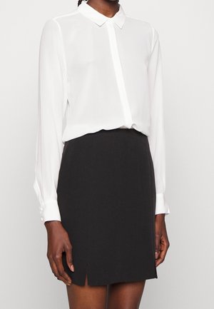 Persona vistiendo una blusa blanca de manga larga con cuello, metida dentro de una falda negra hasta la rodilla con una abertura frontal.