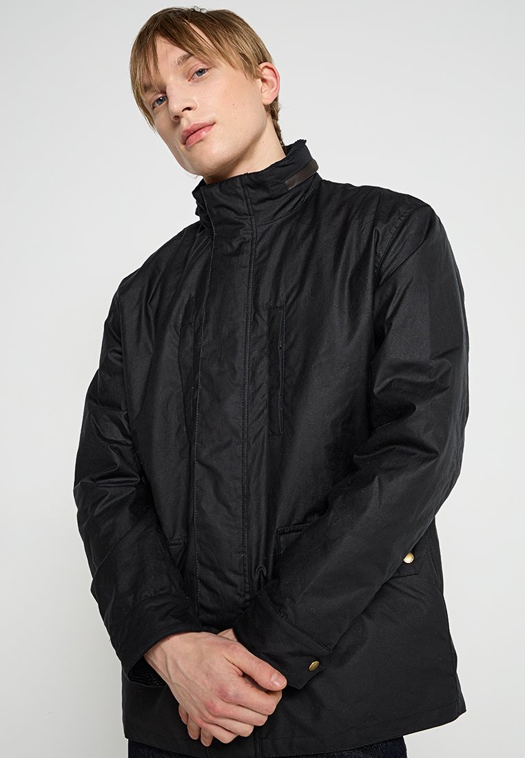 La veste noire présente un col haut, une fermeture à glissière à l'avant et deux poches latérales. Le tissu semble durable, avec une finition mate et des coutures subtiles.