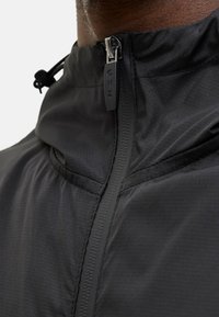 Veste légère noire avec un col montant, fermeture éclair à l'avant avec tirette, et tissu texturé. Comprend des cordons de serrage réglables à la capuche.