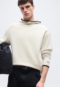 Νέος άνδρας με κρεμ φούτερ με κουκούλα και μαύρο παντελόνι, κρατώντας μια μαύρη τσάντα στο αριστερό του χέρι, μπροστά σε ένα απλό ανοιχτό φόντο.