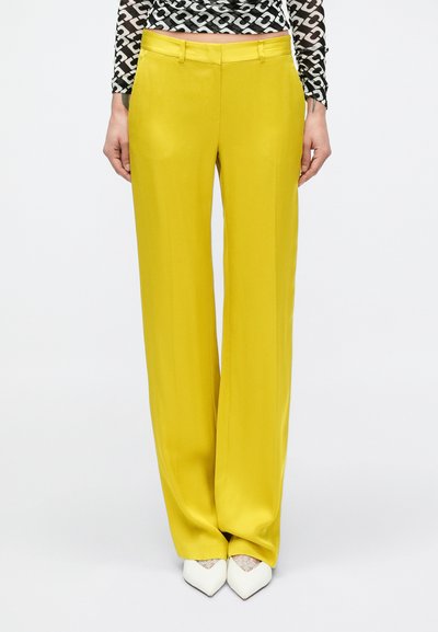 Pantalon jaune à jambes larges avec une texture lisse et des passants pour ceinture. Associé à des talons blancs à bouts pointus pour un contraste.