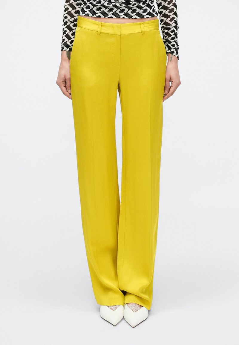 Gele wijde broek met een gladde textuur en riemlussen. Gecombineerd met witte pumps met puntige neus voor contrast.