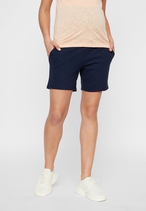 Personne portant un short bleu marine, un haut beige et des baskets blanches, debout les mains dans les poches devant un fond uni.