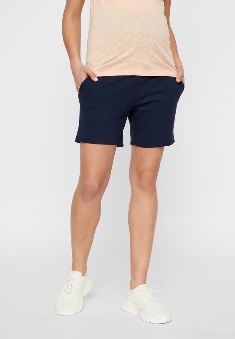Personne portant un short bleu marine, un haut beige et des baskets blanches, debout les mains dans les poches devant un fond uni.