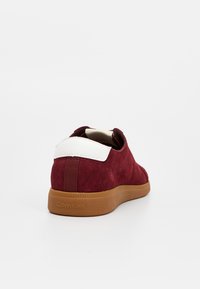 Czerwony zamszowy tenisówka z białym skórzanym paskiem na pięcie i brązową gumową podeszwą. Charakteryzuje się minimalistycznym designem z subtelnym brandingiem na podeszwie.