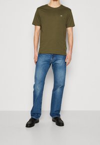 T-shirt a girocollo in cotone verde, abbinato a jeans dritti blu e mocassini neri. La t-shirt presenta un piccolo logo sul petto.