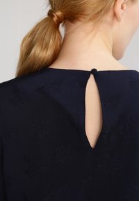 Abito blu marina con un'apertura a chiave sul retro e chiusura con bottone, realizzato in un tessuto textures con motivi floreali e una silhouette semplice ed elegante.