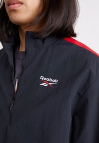 Chaqueta negra con cremallera y acentos rojos en los hombros. Presenta el logo de Reebok en blanco y rojo. Ligera, con textura ripstop.