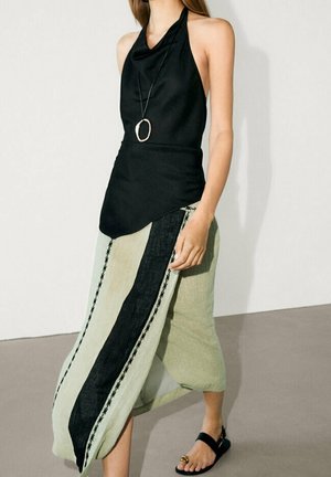 Femme portant un haut noir sans manches à col halter avec pendentif et jupe beige asymétrique avec une bande noire, marchant sur un sol gris en sandales noires.